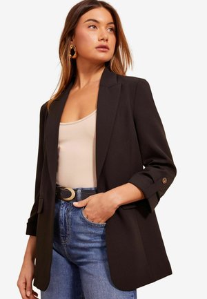 Černý blazer s přizpůsobeným designem, klopovým límcem, ohrnutými rukávy a zlatými knoflíkovými detaily, spárovaný s béžovým topem a modrými džíny.