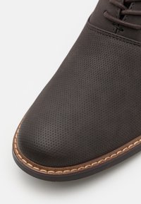 Dark brown perforated leather shoe with a rounded toe, featuring a contrasting stitched edge and smooth rubber sole.