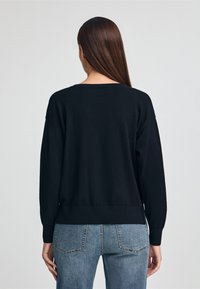 Czarny sweter z dzianiny o luźnym kroju, ze spadniętymi ramionami i ściągaczem na dole. Nosi się go na jasnoniebieskie dżinsy, prezentując prosty, teksturowany wzór.