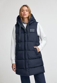 Mörkblå puffväst med huva, med stora quiltade sektioner, sidofickor och en logopatch; tillverkad av ett slätt, isolerat material.