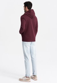 Bordeauxrode hoodie met een trekkoordcapuchon, gemaakt van een zachte katoenmix, gecombineerd met lichtblauwe jeans en beige sneakers met groene accenten.