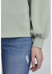 Top de manga larga verde claro con textura y puños fruncidos, combinado con jeans ajustados azules que presentan un parche desgastado.