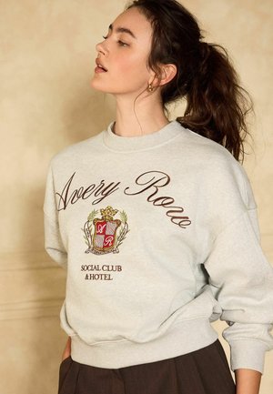 Vrouw met donker haar in een paardenstaart, die een lichtgrijze sweatshirt draagt met "Avery Rose Social Club & Hotel" en embleemtekst aan de voorkant.