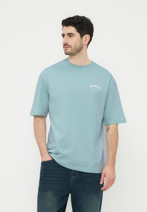 Sötét hajú, szakállas férfi világoskék, túlméretezett pólóban, amin apró fehér feliratok vannak, sötétkék farmernadrágban, kezét zsebre dugva.