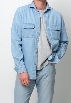 Mand iført lyseblå denim skjorte med knapper over grå t-shirt med matchende lyseblå denim jeans, hånd i lommen, mod ensfarvet baggrund.