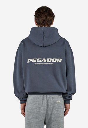 Mężczyzna noszący ciemnoniebieską bluzę z kapturem z napisem "PEGADOR Certified Authentic Streetwear" na plecach oraz jasnoszare spodnie z teksturowanym wzorem.