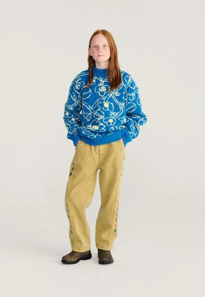 Jeune personne aux longs cheveux roux portant un pull bleu à motifs floraux, un pantalon jaune ample avec des détails colorés, et des bottes marron, debout les mains dans les poches.