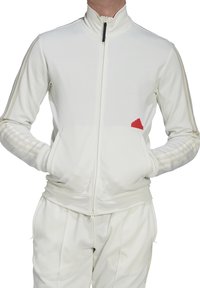 Vit zip-up idrottsjacka med ribbad krage, prydd med subtila tan-ränder på ärmarna och en röd logotypaccent på vänster bröst.