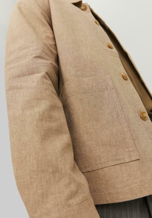 Rudas lininis švarkas su paprastu dizainu, turintis apykaklę, mygtukus ir kišenę šoninėse. Minkšta tekstūra ir pritaikyta forma.