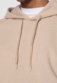 Sudadera beige con capucha y acabado texturizado, con cordones que tienen puntas de plástico y un cuello blanco superpuesto debajo.