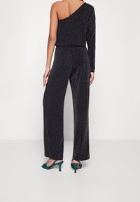 Black jumpsuit with a single long sleeve and an open shoulder, made of sparkly fabric. Paired with teal high-heeled sandals.