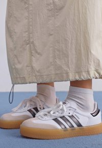 Zapatillas de cuero blancas con rayas negras, suela de goma y detalles texturizados. Arriba, se ve una falda color crema con dobladillo fruncido.