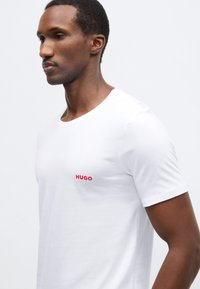 Biele bavlnené tričko s okrúhlym výstrihom, na ľavej strane na hrudi je malý červený nápis "HUGO", hladká textúra a krátke rukávy.