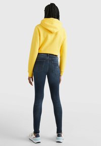 Sudadera amarilla con cordones, combinada con jeans ajustados de color azul oscuro. El atuendo se complementa con zapatillas de color claro con acentos.
