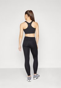 Soutien-gorge de sport noir avec un design dos-nu, associé à un legging noir taille haute. Modèle portant des chaussures de sport grises, vu de dos.