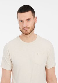 Beżowa koszulka z krótkim rękawem z miękkiego materiału, z małą kieszonką na piersi z metką "PRO TEST". Prosty okrągły dekolt.