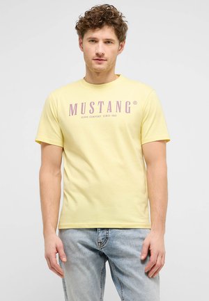 Junger Mann mit lockigem Haar, der ein blassgelbes Mustang-T-Shirt und hellblaue Jeans trägt, steht vor einem schlichten weißen Hintergrund.