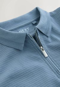 Világoskék pólóing, texturált anyagból, cipzárral és bordázott nyakkal. A címke ezt írja: "NEXT."