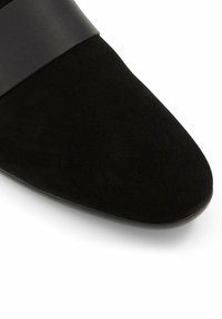 Chaussure plate en daim noir avec bout pointu et détail de bande lisse sur le dessus, présentée sur un fond blanc.