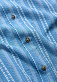 Camisa de tela azul a rayas con rayas blancas verticales, que presenta botones beige. La textura es suave y tiene costuras estándar a lo largo de las uniones.