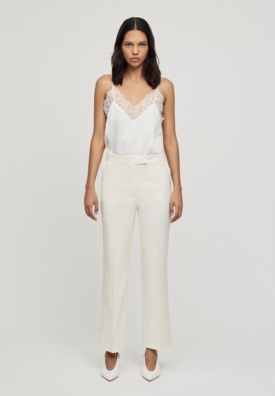 Camisole in satin bianco con bordo in pizzo abbinato a pantaloni beige a vita alta e scarpe bianche con punta affusolata. Sfondo semplice.
