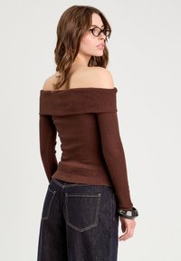 Brun af skulderen ribbet top med lange ærmer, tætsiddende design og tekstureret stof. Båret med mørke denim jeans og et sort armbånd.