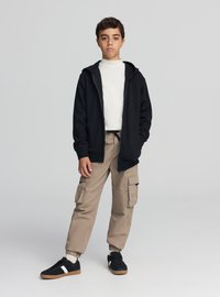 Czarna bluza z kapturem zapinana na zamek, biały golf, beżowe spodnie typu cargo, czarno-białe buty sportowe. Swobodny strój o luźnym kroju z wieloma kieszeniami.