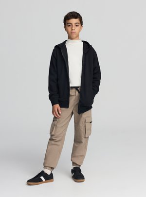 Czarna bluza z kapturem zapinana na zamek, biały golf, beżowe spodnie typu cargo, czarno-białe buty sportowe. Swobodny strój o luźnym kroju z wieloma kieszeniami.