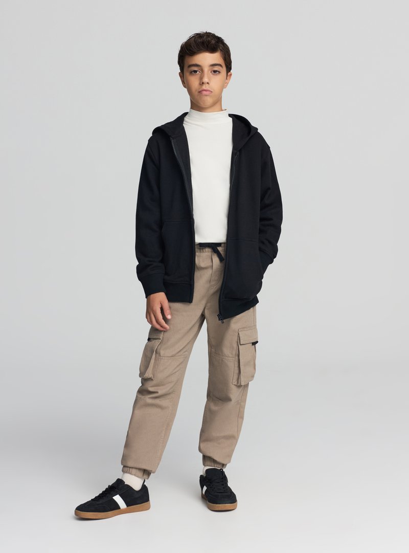 Czarna bluza z kapturem zapinana na zamek, biały golf, beżowe spodnie typu cargo, czarno-białe buty sportowe. Swobodny strój o luźnym kroju z wieloma kieszeniami.