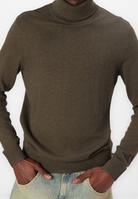 Olijfgroene turtlenecktrui gemaakt van zacht breimateriaal. Fitted ontwerp met geribde manchetten en zoom, gecombineerd met lichte denim jeans.