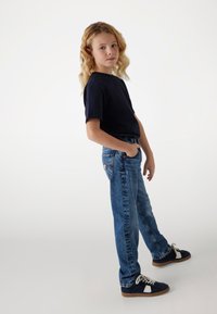 Camicia a maniche corte blu navy, jeans in denim blu con vestibilità classica e sneakers blu navy con dettagli bianchi, in piedi su uno sfondo chiaro.