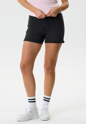 Inimene mustade ülesrullitud äärtega lühikeste pükstega, valgete spordisokidega, millel on kaks musta triipu, valgete tossudega ja heleroosa ülaosaga, käed vöökohal kohandamas.