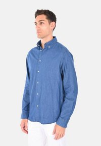Camicia in denim azzurro chiaro con colletto, maniche lunghe e dettagli con bottoni, caratterizzata da una vestibilità comoda e cuciture visibili.