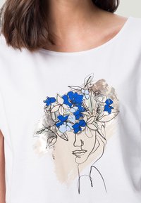 Camiseta blanca con un diseño gráfico de una cara adornada con flores azules y azul claro, contorneada en negro, con acentos dorados.