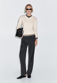 Kvinde iført solbriller, cremet sweater over lag-på-lag toppe, kulgrå vidde-benet bukser, sorte spidse sko, holder en sort læderrygsæk.