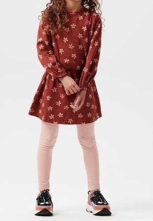 Lapsi punainen kukkakuvioinen mekko päällään, vaaleanpunaiset legginsit ja mustat, vaaleanpunaiset ja valkoiset tennarit jalassa, seisoo yksinkertaisen valkoisen taustan edessä.