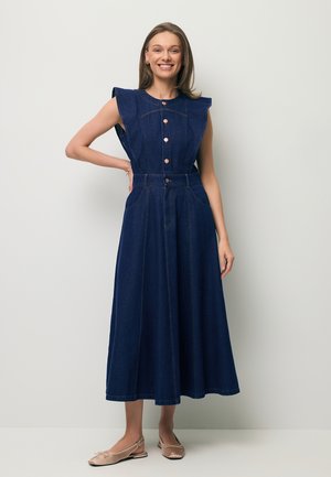 Vrouw die staat en lacht, draagt een mouwloze donkerblauwe denim midi-jurk met knopen en beige ballerina's.