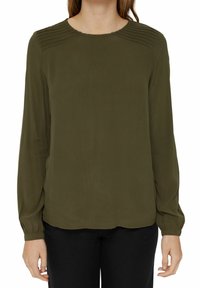 Blusa verde oliva con maniche lunghe, dettaglio sulle spalle plissettato, scollatura tonda e texture liscia. Abbinata a pantaloni neri.