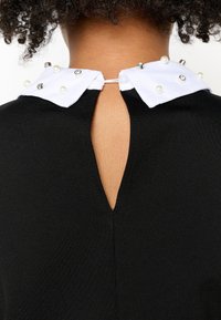 Garb maglia nera con un colletto bianco. Il colletto presenta perle decorative e dettagli metallici, con un'apertura a chiave nella parte posteriore.