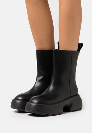 Bottes noires en cuir jusqu'à la cheville, dotées d'une semelle épaisse et texturée et d'un design lisse et épuré avec des languettes sur les côtés.