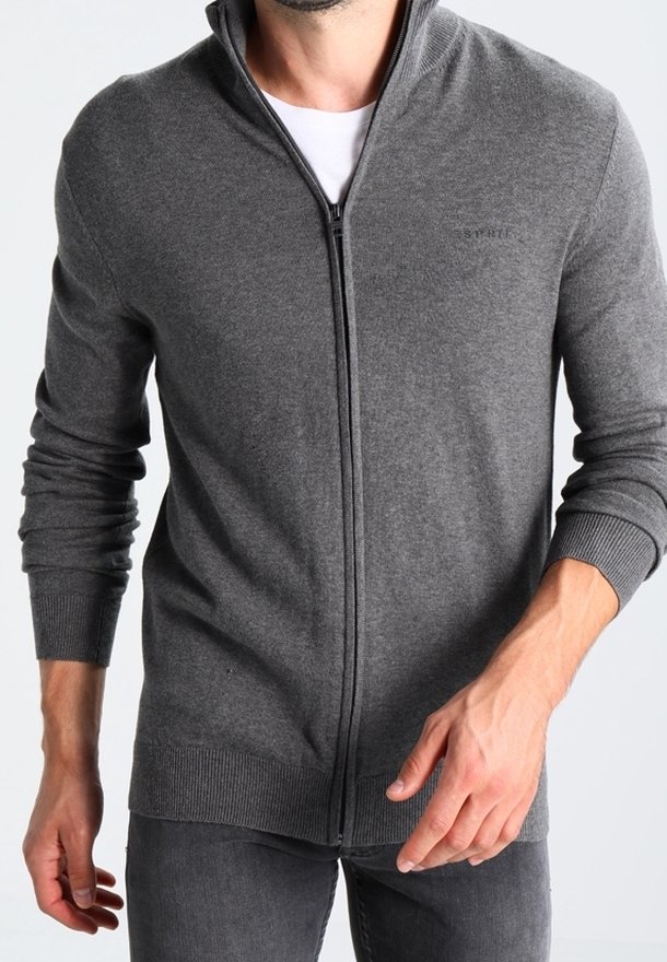 Homme portant un pull zippé gris par-dessus une chemise blanche à manches longues avec des poignets côtelés, associé à un pantalon gris foncé.