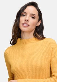 Frau mit welligem dunklem Haar, die einen senfgelben gerippten Pullover trägt und mit einem neutralen Ausdruck vor einem schlichten hellen Hintergrund posiert.