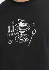 Camiseta negra con un gráfico blanco de un postre en un vaso con una cuchara, junto al texto “OUI S'IL TE PLAÎT” y “-ISOLID.”