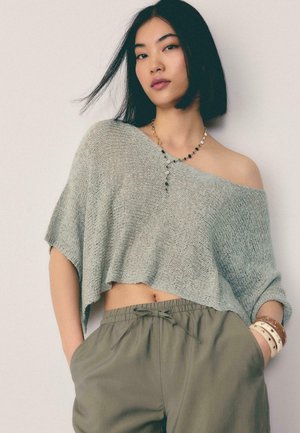 Mladá žena s čiernymi vlasmi, oblečená v šedej pletenej crop top blúzke s odhalenými ramenami, olivových nohaviciach so sťahovacou šnúrkou, vrstvenom náhrdelníku a viacerých náramkoch.