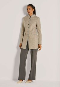 Vrouw in een beige knopenjas, grijze wijde broek en beige puntige hakken, staande tegen een effen lichte achtergrond.