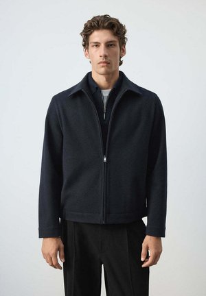 Jeune homme portant une veste zippée bleu marine foncé et un pantalon noir, debout devant un fond gris clair uni.