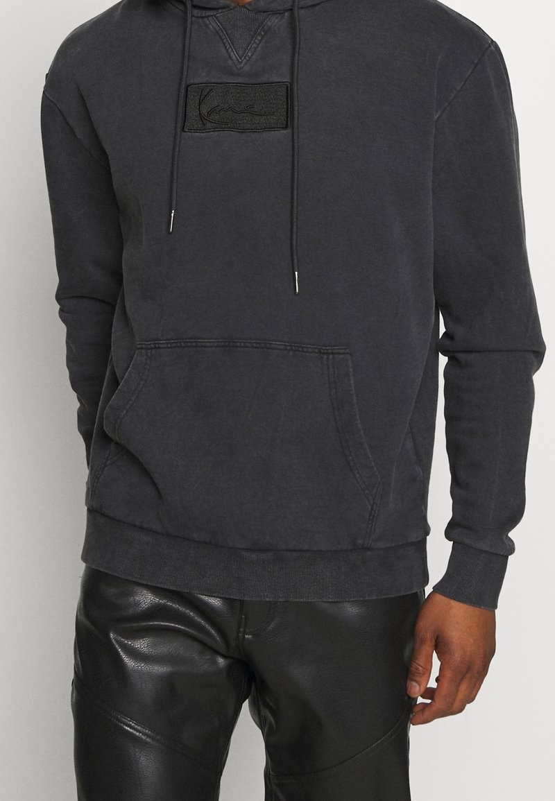 Sudadera capucha gris oscuro con bolsillo delantero, puños acanalados y dobladillo. Presenta un logotipo de parche texturizado negro en el pecho. Textura de tejido suave.