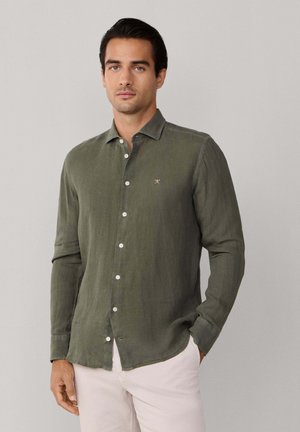 Man draagt een olijfgroene overhemd met lange mouwen en lichtbeige broek, staand tegen een effen lichtgrijze achtergrond.