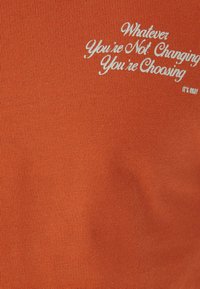 Orange bomulds t-shirt med hvid broderet tekst med sætningen: "Hvad end du ikke ændrer, vælger du DET ER OKAY."