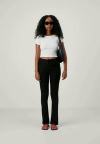 Γυναίκα με σγουρά μαλλιά που φορά μαύρα γυαλιά ηλίου, λευκό cropped t-shirt, μαύρα παντελόνια, κόκκινες σαγιονάρες και κρατά μια μαύρη τσάντα ώμου, στέκεται μπροστά σε απλή φόντο.